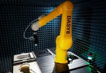 Un bras robotique jaune étiqueté "DASY8" fait une démonstration de robotisation dans une chambre anéchoïque, entourée de panneaux de mousse absorbant le son et d'équipements. Un petit voyant lumineux est visible sur le côté droit de la table.