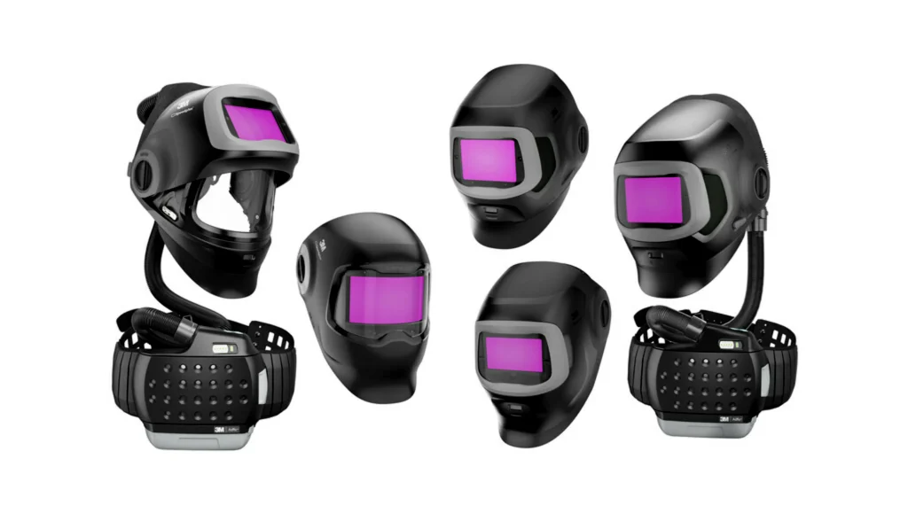Six casques de soudage Speedglas noirs avec des visières teintées en violet sont disposés en deux rangées. Certains casques, véritables pionniers de la protection de soudage, sont équipés de respirateurs et de tuyaux. L'arrière-plan est blanc.