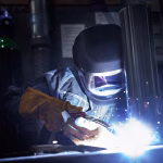 Une personne portant des gants de protection et un casque de soudage Speedglas utilise une torche de soudage, produisant de vives étincelles et de la lumière, dans un atelier industriel axé sur la protection de soudage.