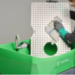 Une personne portant des gants essuie un panneau en plastique perforé avec un chiffon. Le panneau est posé sur un conteneur vert étiqueté "Mewa".