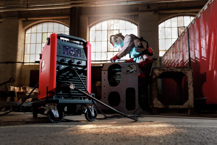 Au premier plan d'un atelier industriel, une machine à souder rouge sur roues est fixée à des câbles. À l'arrière-plan, une personne en tenue de sécurité travaille sur une grande pièce métallique près de hautes fenêtres.