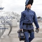 Un homme portant un équipement de protection pour le soudage transporte un équipement de soudage portable CORE à l'extérieur, près d'un équipement industriel et de piles de roches. Le texte se lit comme suit : "Böhler, soudage par voestalpine. Allez souder ! Avec Core. Une nouvelle ère dans l'équipement de soudage pour les professionnels.