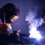 Une personne portant un casque de protection utilise un outil de soudage sur du métal. Des étincelles et de la fumée s'élèvent de la zone de travail lors d'un webinaire de l'Institut de Soudure.
