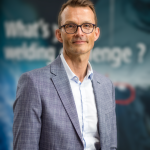 Un homme portant des lunettes et un costume gris clair avec une chemise blanche se tient devant un fond flou avec un texte partiellement visible.