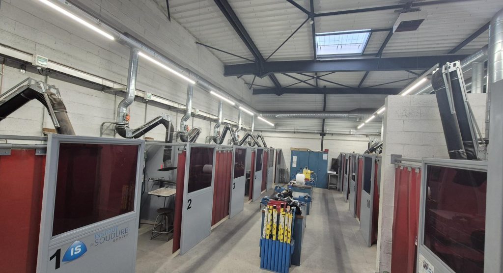 Une rangée de cabines de soudage numérotées avec des cloisons rouges borde les deux côtés d'un atelier industriel. Le plafond est haut, avec des poutres apparentes, des conduits de ventilation et des puits de lumière. Le matériel et les fournitures sont visibles dans l'allée centrale.