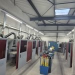 Une rangée de cabines de soudage numérotées avec des cloisons rouges borde les deux côtés d'un atelier industriel. Le plafond est haut, avec des poutres apparentes, des conduits de ventilation et des puits de lumière. Le matériel et les fournitures sont visibles dans l'allée centrale.