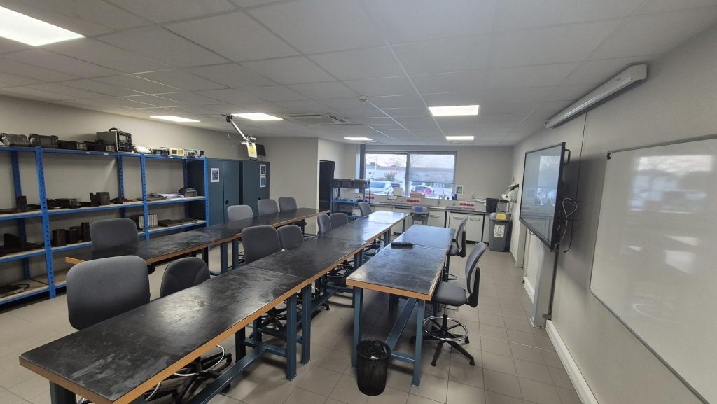 Une salle de classe avec de longues tables noires et des chaises grises disposées en deux rangées, des étagères bleues avec du matériel le long du mur de gauche, une grande fenêtre au fond, un tableau blanc et un écran numérique sur le mur de droite.