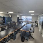 Une salle de classe avec de longues tables noires et des chaises grises disposées en deux rangées, des étagères bleues avec du matériel le long du mur de gauche, une grande fenêtre au fond, un tableau blanc et un écran numérique sur le mur de droite.