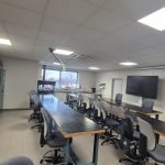 Une salle de classe moderne avec de longues tables noires, des chaises grises sur roulettes, des étagères métalliques bleues contenant du matériel, de grandes fenêtres, un écran mural et un tableau blanc éclairé par des plafonniers.