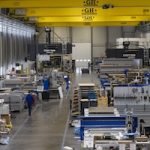 Intérieur d'une grande usine industrielle avec diverses machines et équipements, des ponts roulants jaunes et quelques ouvriers en uniforme bleu marchant et travaillant le long de larges allées. L'espace est très éclairé et les plafonds sont hauts.