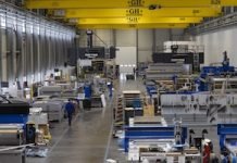 Intérieur d'une grande usine industrielle avec diverses machines et équipements, des ponts roulants jaunes et quelques ouvriers en uniforme bleu marchant et travaillant le long de larges allées. L'espace est très éclairé et les plafonds sont hauts.