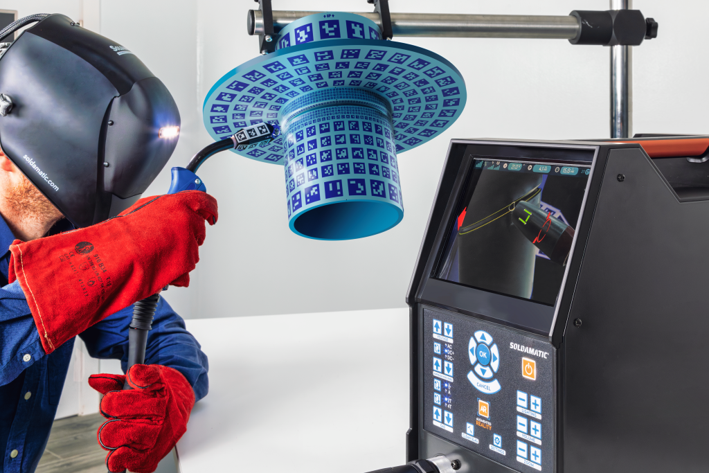 Une personne portant un casque de soudure et des gants rouges tient une torche de soudage près d'un objet cylindrique bleu avec une surface à motifs, tandis qu'une machine située à proximité affiche des données connexes et une vue rapprochée sur son écran.