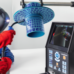 Une personne portant un casque de soudure et des gants rouges tient une torche de soudage près d'un objet cylindrique bleu avec une surface à motifs, tandis qu'une machine située à proximité affiche des données connexes et une vue rapprochée sur son écran.