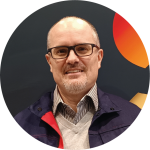 Un homme d'âge moyen avec des lunettes, une courte barbe et une ligne de cheveux fuyante sourit. Vêtu d'une chemise à col, d'un pull et d'une veste, il se tient devant un arrière-plan sombre orné de formes orange et rouges, qui reflètent les efforts déployés pour reconstruire l'industrie.