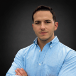 Un homme aux cheveux courts et bruns, vêtu d'une chemise à boutons bleu clair, se tient debout, les bras croisés, devant un fond uni et sombre. Il incarne l'assurance que l'on retrouve souvent dans l'industrie ou chez ceux qui suivent une formation de soudeur.