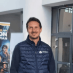 Un homme portant un gilet matelassé bleu se tient à l'intérieur, près d'une porte vitrée, avec une affiche de film représentant plusieurs visages et le mot "MAN" partiellement visible à l'arrière-plan, suggérant un lien avec la formation dans l'industrie.