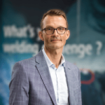 Un homme aux cheveux courts et aux lunettes, vêtu d'une veste de costume gris clair et d'une chemise blanche, se tient devant un arrière-plan flou sur lequel figure le texte "What's your wedding challenge", qui met en lumière l'innovation dans l'industrie.