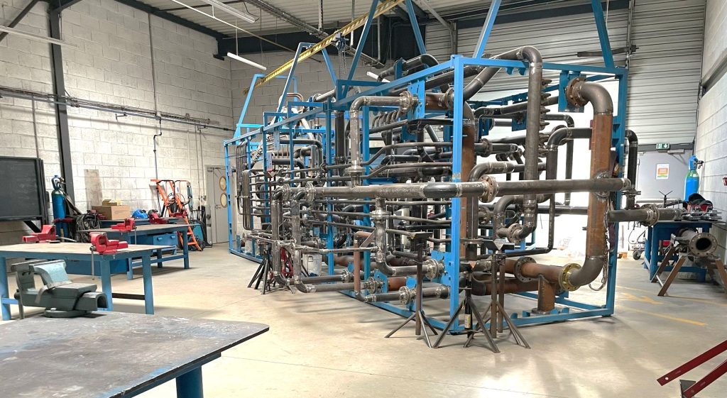 Un grand cadre métallique bleu soutient un réseau complexe de tuyaux métalliques dans un atelier industriel. Des établis, des outils et des équipements sont visibles dans cette pièce spacieuse et bien éclairée.