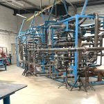 Un grand cadre métallique bleu soutient un réseau complexe de tuyaux métalliques dans un atelier industriel. Des établis, des outils et des équipements sont visibles dans cette pièce spacieuse et bien éclairée.