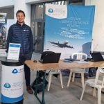 Un homme portant un gilet bleu se tient à côté d'un stand de l'"Institut de Soudure" avec une grande bannière représentant un avion, des tables avec des ordinateurs portables et des documents d'information lors d'un événement en intérieur.