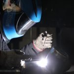 Une personne portant des gants de protection et un casque de soudure bleu soude des tuyaux métalliques, une lumière vive étant visible au point de soudure dans un environnement faiblement éclairé.