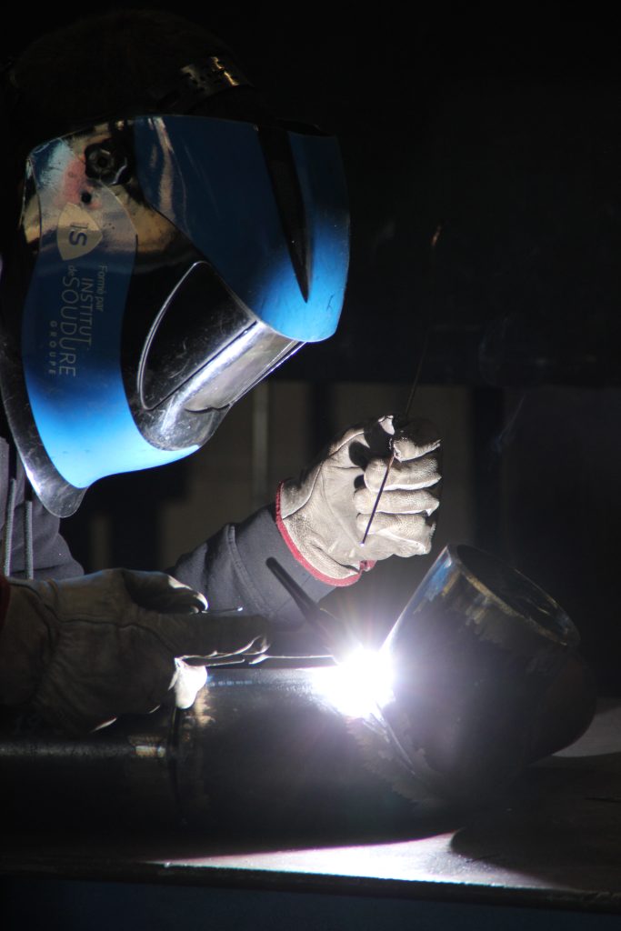 Une personne portant des gants de protection et un casque de soudure bleu soude des tuyaux métalliques, une lumière vive étant visible au point de soudure dans un environnement faiblement éclairé.