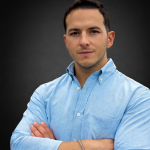 Un homme aux cheveux courts et foncés, portant une chemise bleu clair boutonnée, se tient debout, les bras croisés, sur un fond uni et sombre. Il regarde directement la caméra avec une expression neutre.