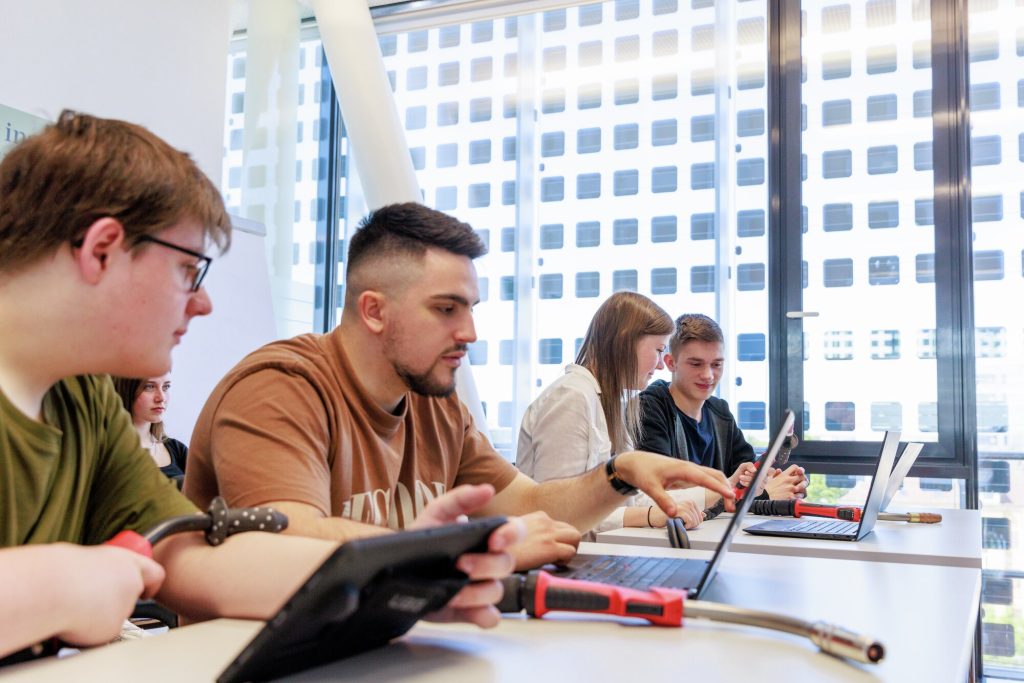 Quatre étudiants sont assis à une table, utilisant des ordinateurs portables et tenant des outils, travaillant ensemble dans une salle lumineuse avec de grandes fenêtres montrant un bâtiment moderne à l'extérieur.