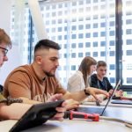 Quatre étudiants sont assis à une table, utilisant des ordinateurs portables et tenant des outils, travaillant ensemble dans une salle lumineuse avec de grandes fenêtres montrant un bâtiment moderne à l'extérieur.