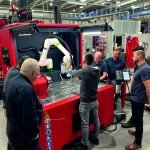 Cinq personnes se tiennent autour d'une grande machine industrielle rouge dotée d'un bras robotisé dans une usine. Une personne fait fonctionner la machine tandis que les autres observent. Diverses commandes et étiquettes de sécurité sont visibles.