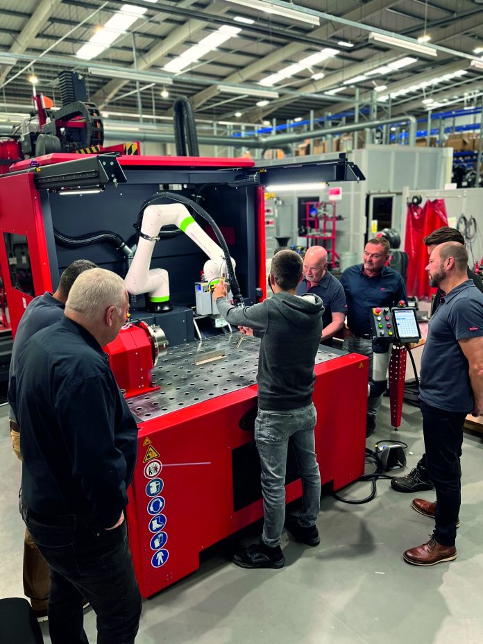 Cinq personnes se tiennent autour d'une grande machine industrielle rouge dotée d'un bras robotisé dans une usine. Une personne fait fonctionner la machine tandis que les autres observent. Diverses commandes et étiquettes de sécurité sont visibles.
