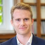 Un homme aux cheveux blonds courts, vêtu d'une veste de costume marine et d'une chemise blanche, se tient à l'intérieur devant un arrière-plan flou composé d'étagères de livres et d'une fenêtre, suggérant un cadre axé sur la formation ou l'entraînement dans le secteur de l'industrie.