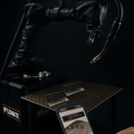 Une personne tient un smartphone affichant une application de soudage devant un robot de soudage automatisé et une table de travail en métal avec des pièces métalliques, soulignant l'engagement de WELDMATE pour la formation des soudeurs dans l'industrie moderne.