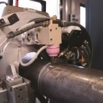 Vue rapprochée d'une machine industrielle soudant ou travaillant sur la surface d'un grand tuyau métallique, montrant des pièces mécaniques, des câbles et une buse entrant en contact avec le tuyau.