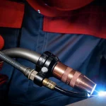 Une personne portant des gants et des vêtements de protection utilise une torche de soudage, produisant un arc lumineux bleu sur une surface métallique.