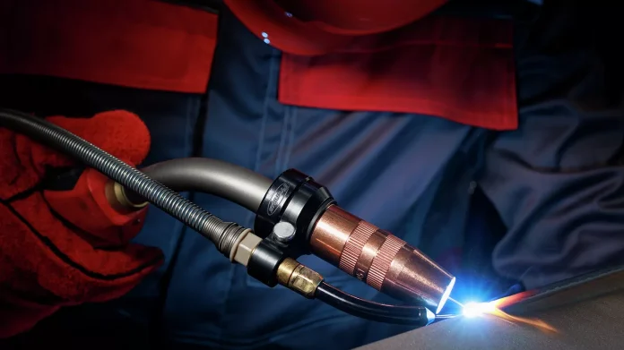 Une personne portant des gants et des vêtements de protection utilise une torche de soudage, produisant un arc lumineux bleu sur une surface métallique.