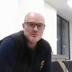 Un homme à lunettes et à la ligne de cheveux fuyante est assis à une table, à l'intérieur, et discute peut-être de la formation des soudeurs dans l'industrie. Il porte un gilet sombre sur une chemise, avec de grandes fenêtres et des murs clairs en arrière-plan.