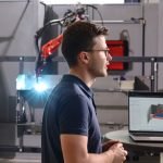 Un homme portant des lunettes se tient à côté d'un ordinateur portable affichant un modèle en 3D, avec en arrière-plan un équipement industriel et un bras robotisé. La scène semble se dérouler dans une usine moderne de fabrication ou d'ingénierie.