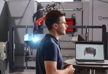 Logiciel ARP d’ArcNC : une avancée majeure en soudage robotisé Un homme portant des lunettes se tient à côté d'un ordinateur portable affichant un modèle en 3D, avec en arrière-plan un équipement industriel et un bras robotisé. La scène semble se dérouler dans une usine moderne de fabrication ou d'ingénierie.