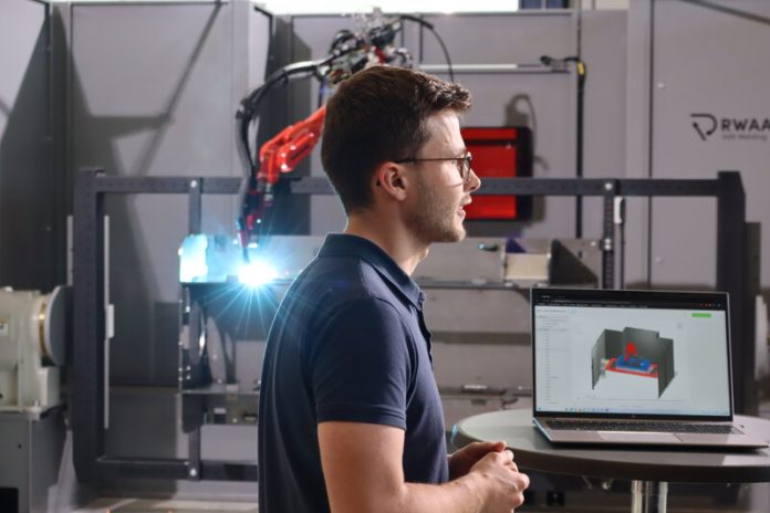 Un homme portant des lunettes se tient à côté d'un ordinateur portable affichant un modèle en 3D, avec en arrière-plan un équipement industriel et un bras robotisé. La scène semble se dérouler dans une usine moderne de fabrication ou d'ingénierie.