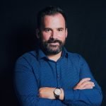 Un homme aux cheveux et à la barbe foncés se tient sur un fond noir, vêtu d'une chemise bleue boutonnée à petits points blancs. Il a les bras croisés et regarde la caméra. Il porte une montre au poignet gauche.