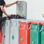 Une personne dépose un objet dans une poubelle grise à couvercle surélevé. Plusieurs bacs fermés, gris, rouges et verts, sont alignés contre un mur dans un environnement intérieur propre. Des tuyaux et des vannes sont visibles sur le mur.