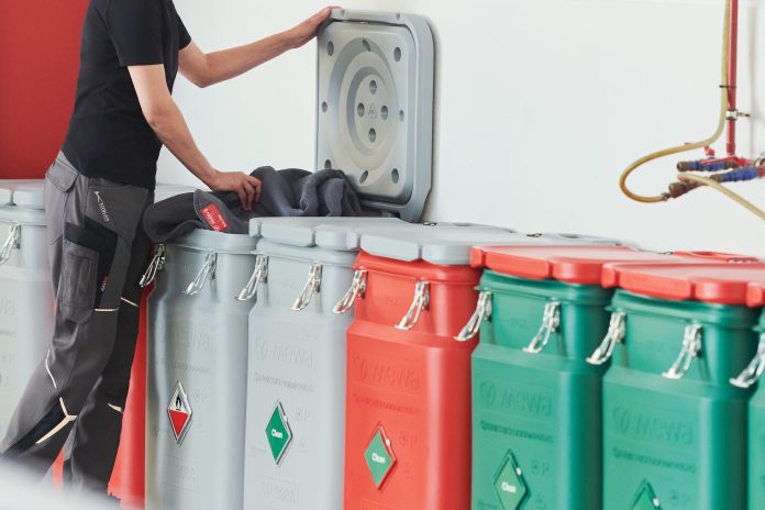 Une personne dépose un objet dans une poubelle grise à couvercle surélevé. Plusieurs bacs fermés, gris, rouges et verts, sont alignés contre un mur dans un environnement intérieur propre. Des tuyaux et des vannes sont visibles sur le mur.