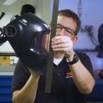 Une personne portant des lunettes tient un casque de soudure à la verticale dans une main et mesure sa hauteur à l'aide d'une règle en métal. L'arrière-plan montre des équipements et des outils d'atelier.