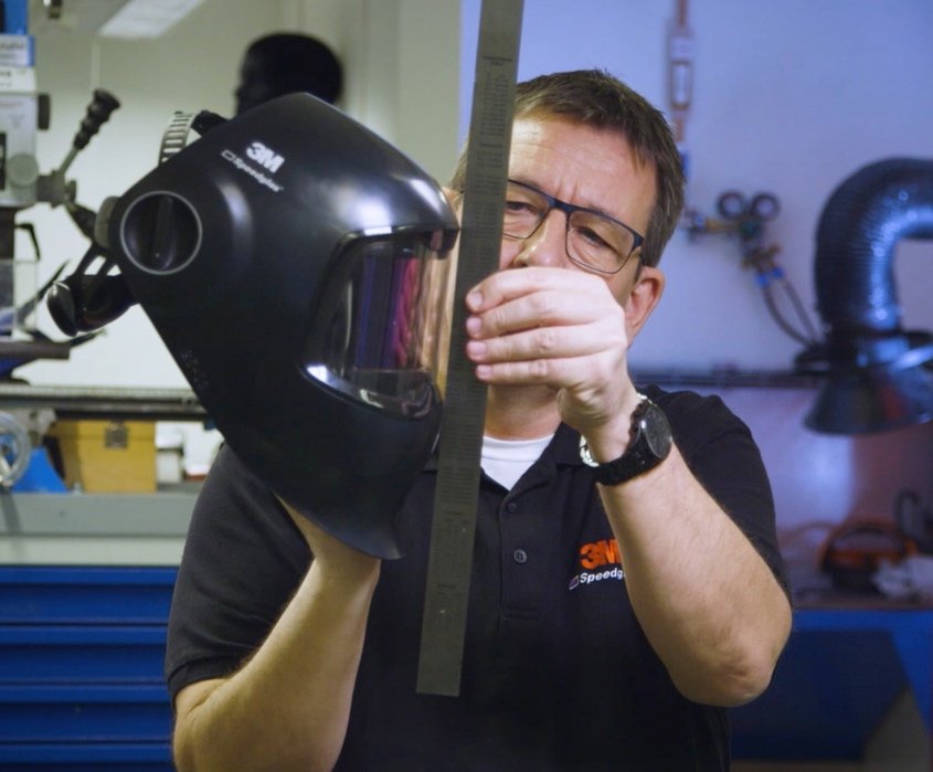 Une personne portant des lunettes tient un casque de soudure à la verticale dans une main et mesure sa hauteur à l'aide d'une règle en métal. L'arrière-plan montre des équipements et des outils d'atelier.