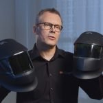 Un homme portant des lunettes et une chemise noire tient deux casques de soudure noirs, un dans chaque main, debout à l'intérieur devant une fenêtre aux rideaux blancs.