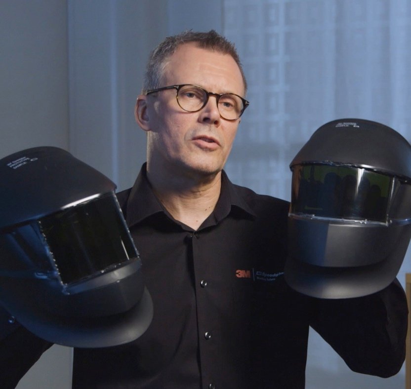 Un homme portant des lunettes et une chemise noire tient deux casques de soudure noirs, un dans chaque main, debout à l'intérieur devant une fenêtre aux rideaux blancs.