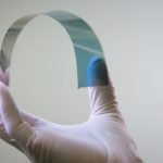 Une main munie d'un gant en latex blanc tient une feuille mince, flexible et semi-transparente sur un fond clair uni.