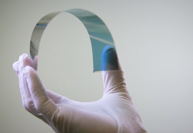 Une main munie d'un gant en latex blanc tient une feuille mince, flexible et semi-transparente sur un fond clair uni.