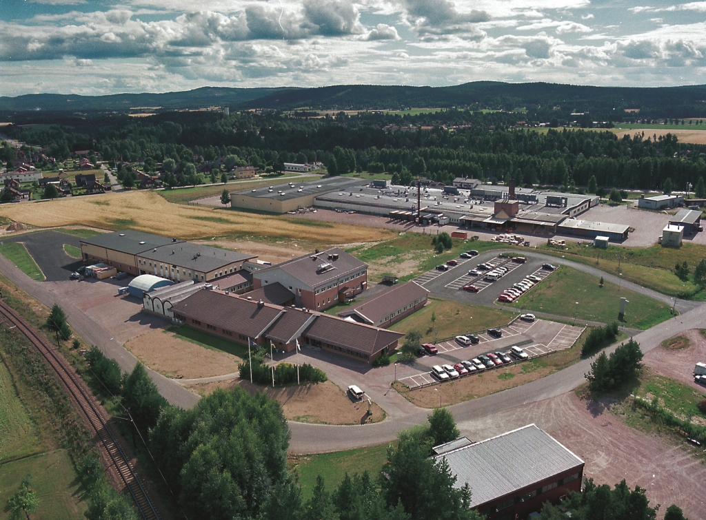 Vue aérienne d'un grand complexe immobilier avec plusieurs parkings, entouré d'arbres, de champs et de collines lointaines, sous un ciel partiellement nuageux. Plusieurs voitures sont garées et une voie ferrée longe le bord inférieur.